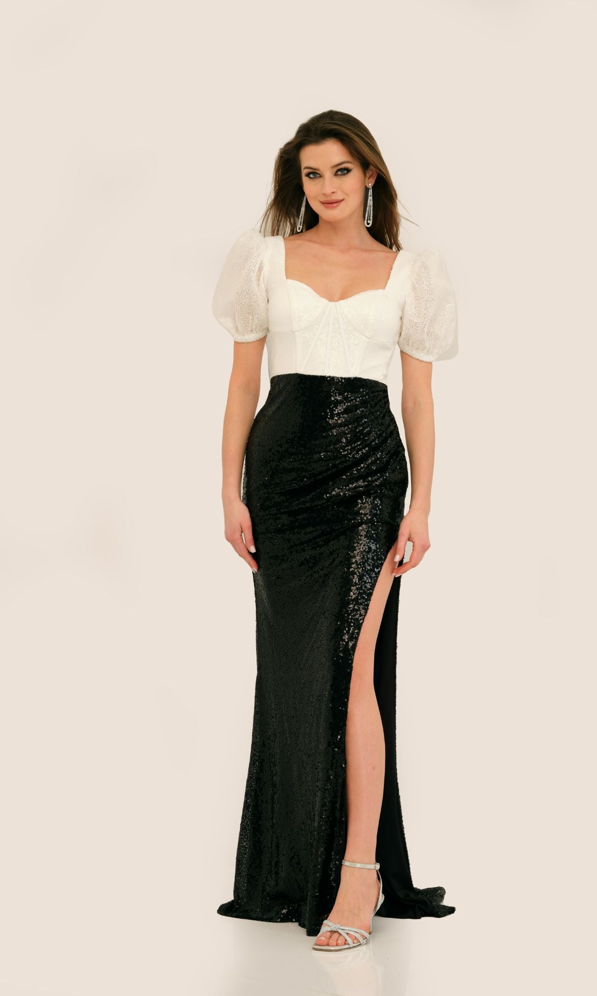 A woman models the Long Formal Dress 11328 by Dave and Johnny—a black sequin floor-length gown with a thigh-high slit—paired with silver heels and drop earrings, set against a plain background.